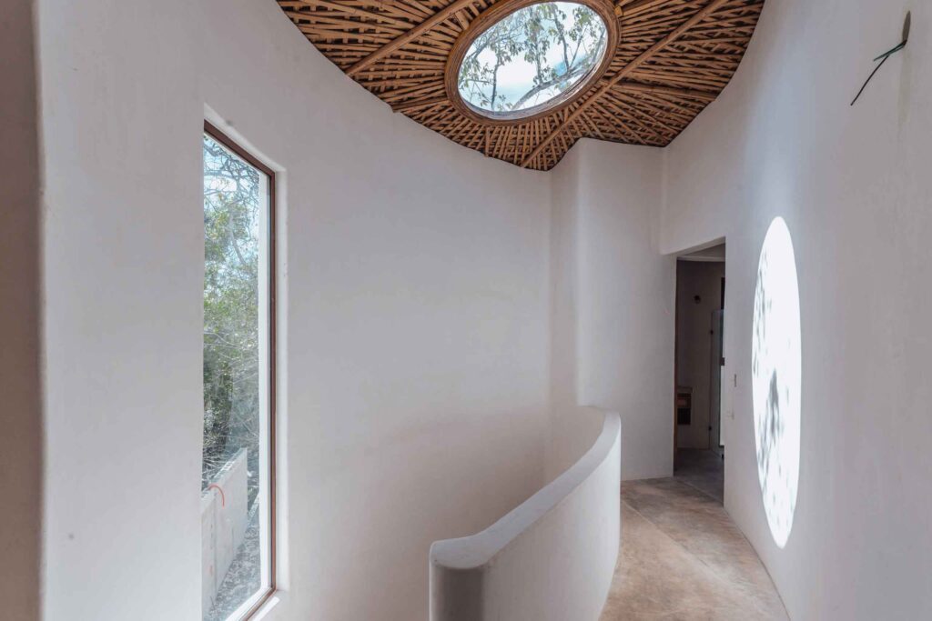 A curved hallway with white walls, a round skylight with a woven wooden pattern, a large window, and sunlight casting a circular shadow on the wall. Apartments and villas for rent in Tulum