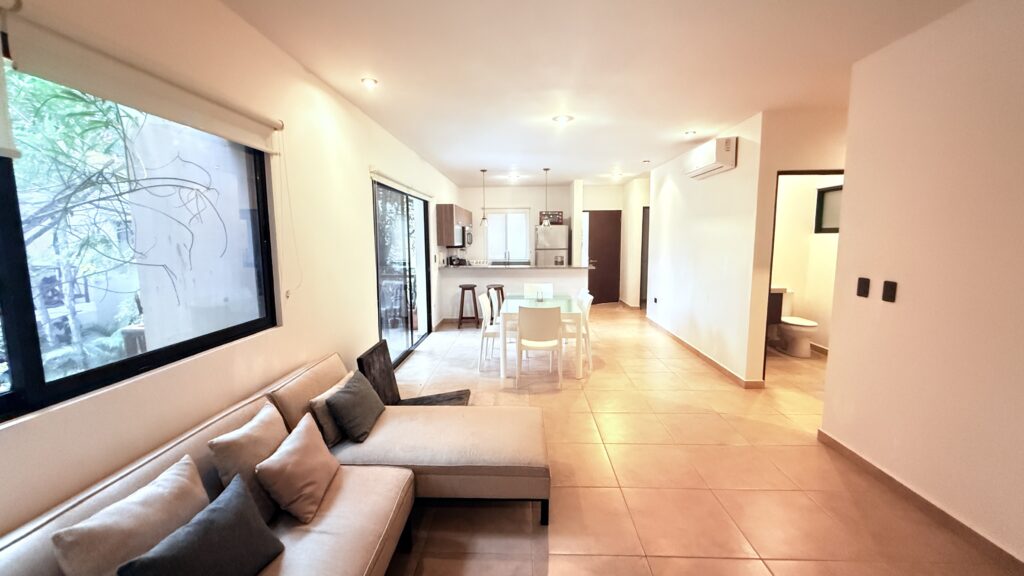 Bright, modern living room with a sectional sofa, dining table, bar stools, open kitchen, tiled floor, and large windows. Apartments and villas for rent in Tulum