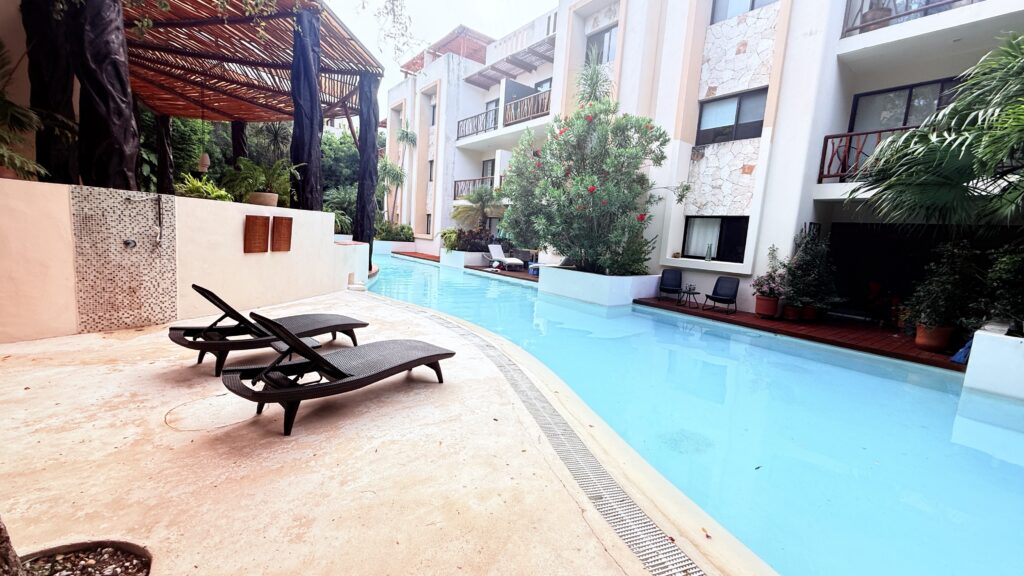 Two black lounge chairs sit beside a curved outdoor pool, adjacent to a white apartment building with balconies and lush green plants. Apartments and villas for rent in Tulum