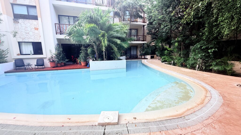 Outdoor swimming pool with curved edges, surrounded by tropical plants and a multi-story building with balconies and windows. Apartments and villas for rent in Tulum